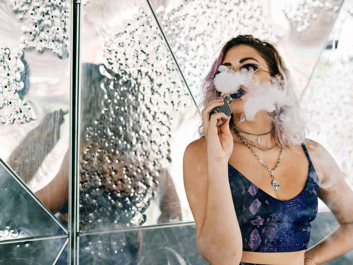 woman blowing a cloud of smoke after using a cbd vape against a mirror
