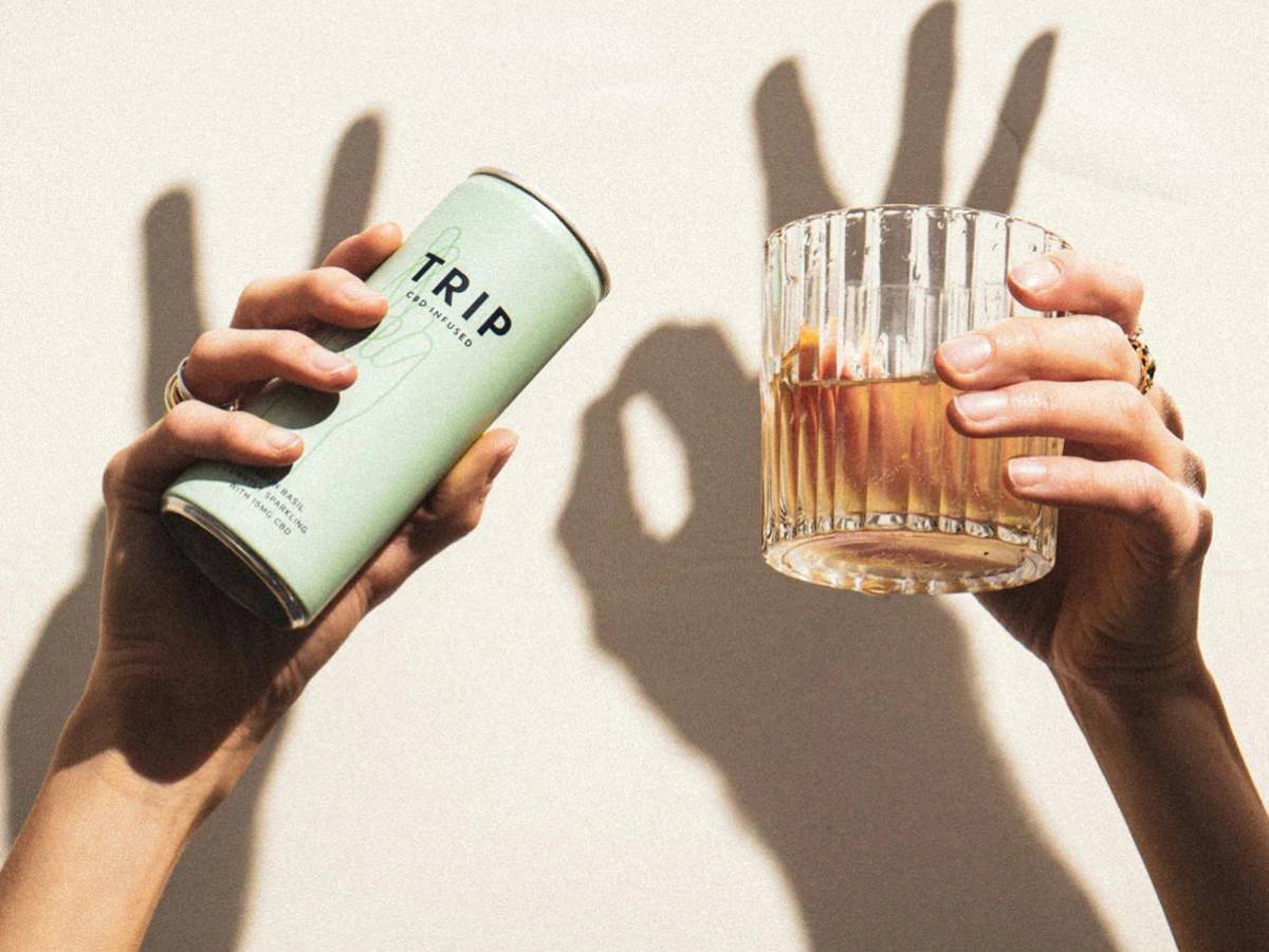 hands holding up a tin of trip cbd elderflower mint drink and a glass of it with an orange wedge. behind it, shadows make a peace sign and 'ok' sign