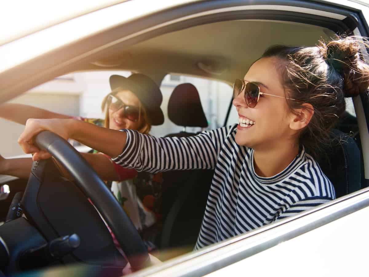 two women smiling and driving in a car with sunglasses on and sunbeams through the window