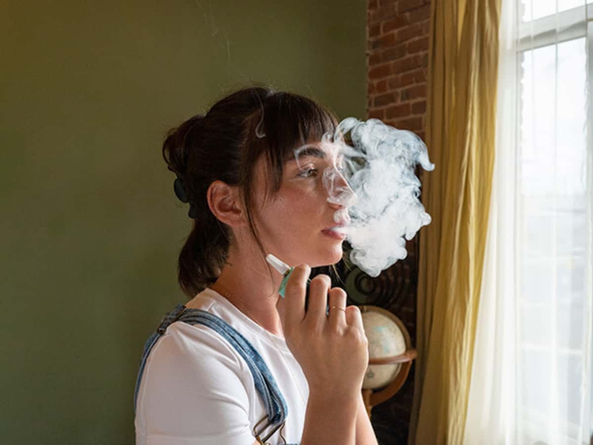 A woman at a window exhaling a cloud of smoke from a ccell battery pen and cbd cartridge