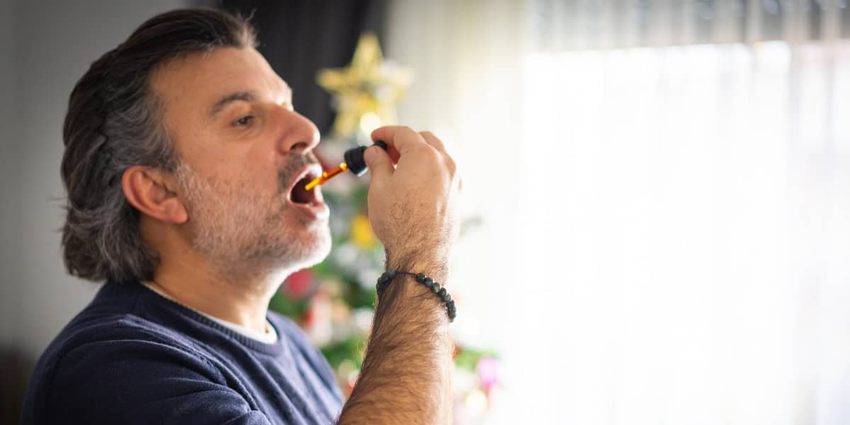 Man administering CBD oil from a pipette with Christmas tree and backlit lights from the curtains