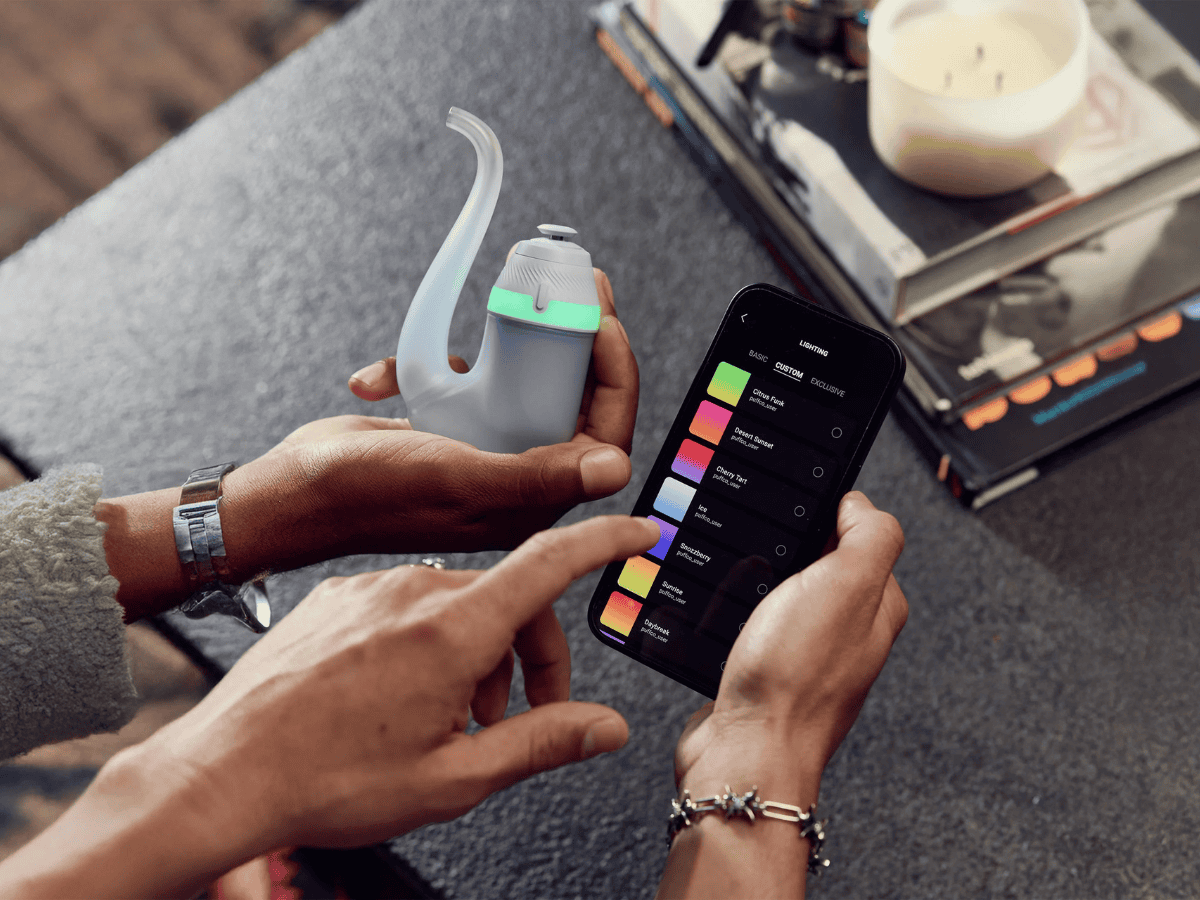 Puffco Proxy Dab Pen and app in use. One had holding the device and another person's two hands holding and working with the app, table background