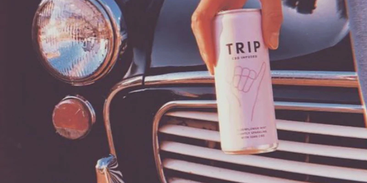 hand holding a can of TRIP CBD drink in front of the grill of a black car